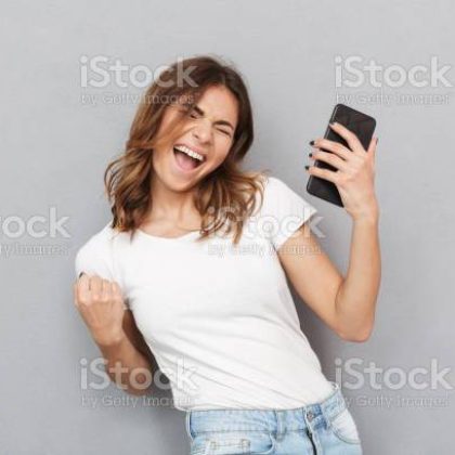 Portrait of a satisfied young woman holding mobile phone and celebrating over gray background